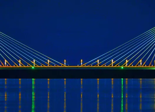 A cable-stayed bridge illuminated at night. The bridge has two towers with cables extending to the road deck. The bridge is reflected in the water below.